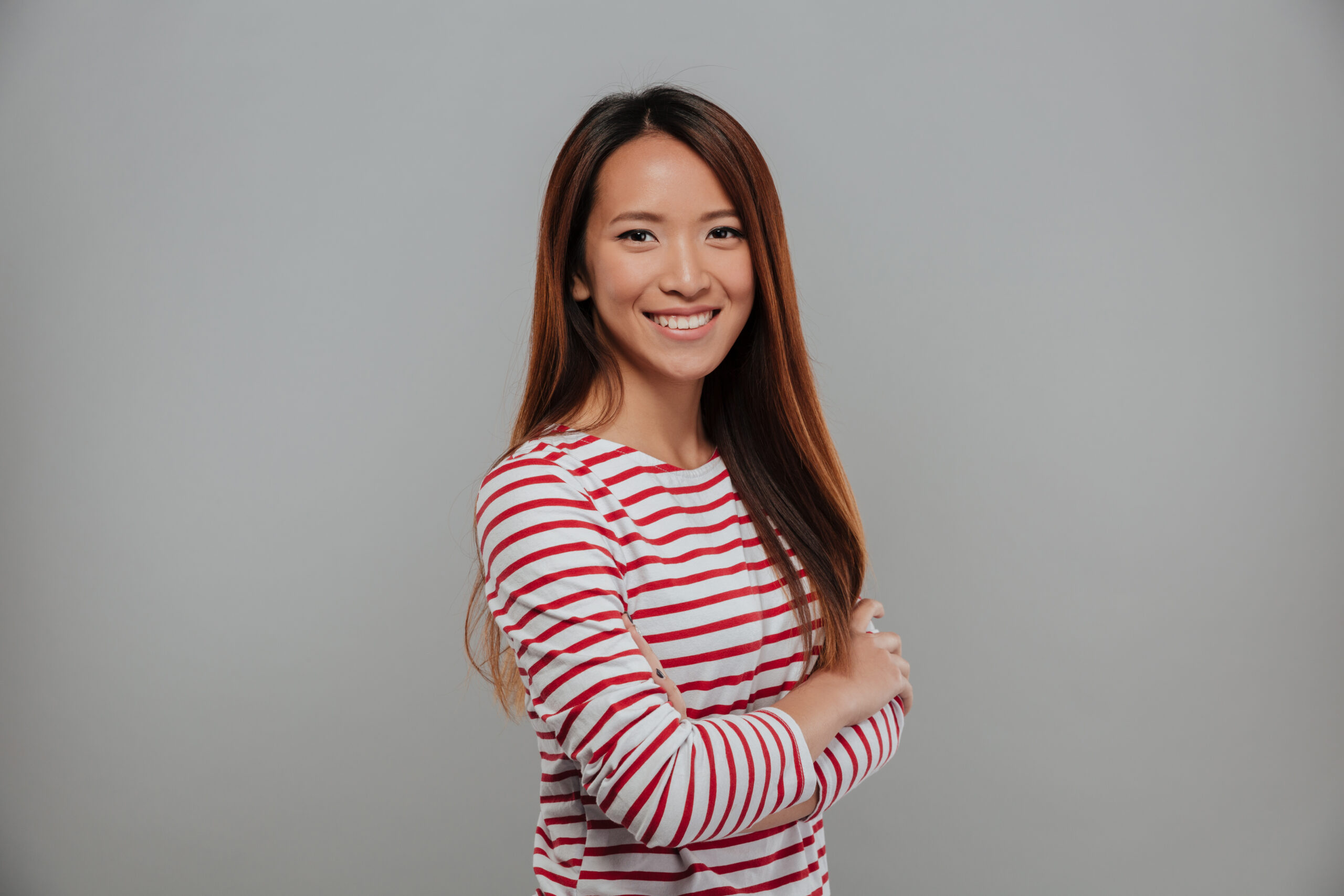Side view of smiling asian woman in sweater posing with crossed arms and looking at the camera over gray background